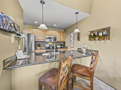 A kitchen with wooden cabinets, stainless steel appliances, a granite countertop, two wooden barstools, and hanging pendant lights. A wine rack is mounted on the wall above the counter.