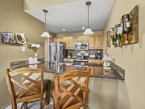 Modern kitchen with wooden cabinets, stainless steel appliances, granite countertops, and a small breakfast bar with two wooden chairs. Wall-mounted racks hold mail and wine bottles.