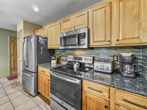 A kitchen with wooden cabinets, stainless steel refrigerator, microwave, electric stove, granite countertops, black and blue tile backsplash, toaster, coffee maker, and a knife block.