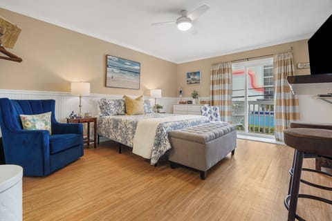 Your spacious bedroom features coastal charm with beach-themed art, a plush blue armchair, and a private balcony overlooking the pool.