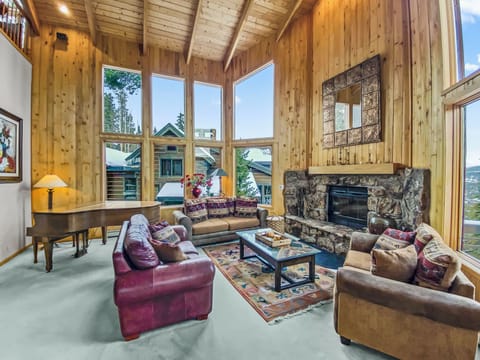 Spacious living room with large windows, wooden walls, vaulted ceiling, stone fireplace, sofas, a piano, glass coffee table, and a patterned area rug. Snowy landscape visible through the windows.