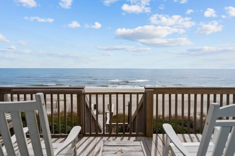 White deck chairs overlook a wooden railing, beach walkway, sandy shore, gentle Atlantic waves, and blue sky, perfect for an Oak Island, NC oceanfront vacation rental.