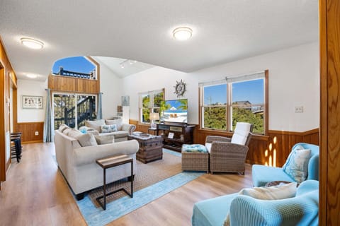The spacious living area features high ceilings, bright natural light, and coastal-inspired décor, creating a warm and inviting place to relax around the flat-screen TV.