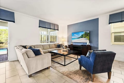 Spacious living room with cream sectional seating, a large flat-screen TV, coastal sailboat accents, and a sunny glimpse of the pool.