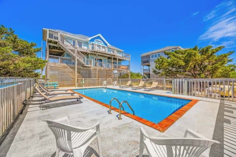 Beachside vacation rental complex featuring a private pool, multiple sun loungers, and elevated oceanfront homes surrounded by coastal greenery under clear blue skies.