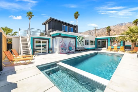 Sunny pool and spa with wide deck, colorful egg chairs, rooftop terrace, and snow-capped mountain views behind palm trees