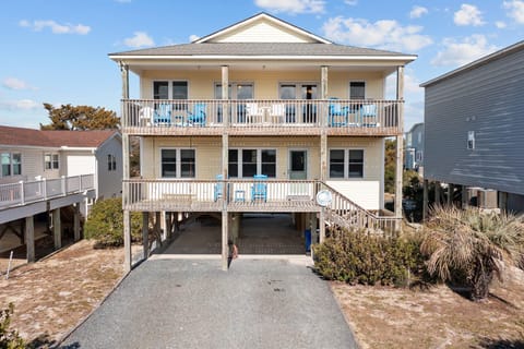 Elevated coastal home with two covered decks, outdoor seating, wood railings, stairs, covered parking, gravel driveway, shrubs, and blue sky.