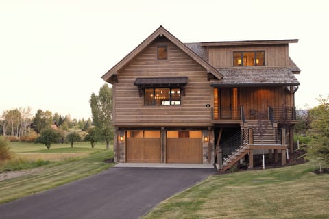 Your stunning cedar retreat awaits with warm lights glowing through expansive windows, inviting you to unwind in this beautifully crafted mountain home surrounded by rolling greens.