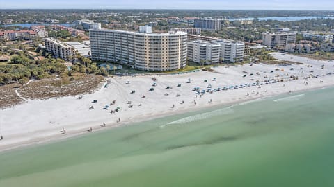 Aerial view of a coastal city featuring a wide sandy beach, ocean waves, beach chairs, umbrellas, and several mid-rise buildings and resorts.