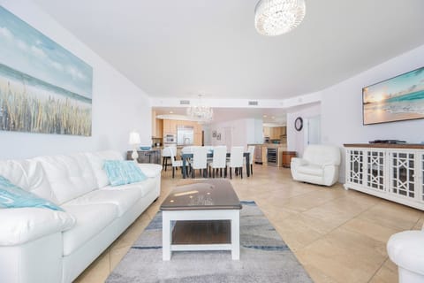 Bright living area featuring a white leather sofas with two queen sleepers, elegant chandelier, and a view into the kitchen