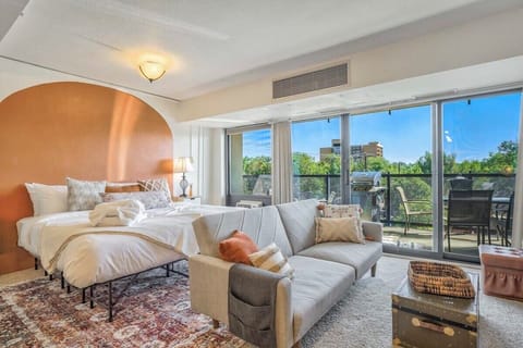 Sunlight pours through wide windows onto a styled bed and cozy seating, creating a calm spot to stretch out with a book or ease into the morning. Warm tones, layered textures, and soft lighting make the room feel inviting from the moment you arrive.