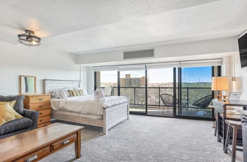 Sunlit living room with plush seating opens to sliding glass doors and a private balcony, inviting slow mornings and evening wind-downs while city buildings and distant hills frame the view.