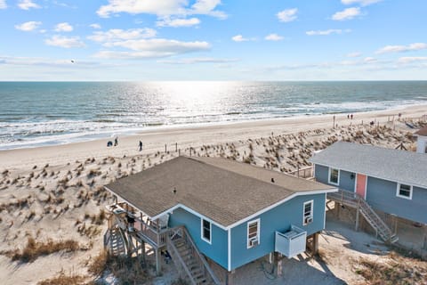 Blue coastal home on dunes with oceanfront deck, beach access, sandy shoreline, sea oats, waves, wide water views, and bright Oak Island, NC sky.