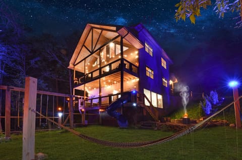 Rustic cabin with illuminated decks, a slide, and a fire pit under a starry night sky.