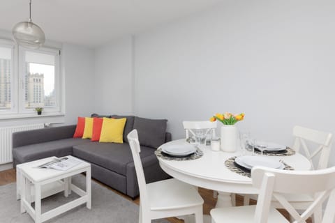 A cozy living room with a gray sofa and a small white round table. The sofa has colorful pillows, and the table is set with a small plant and a book. The room is compact and bright.