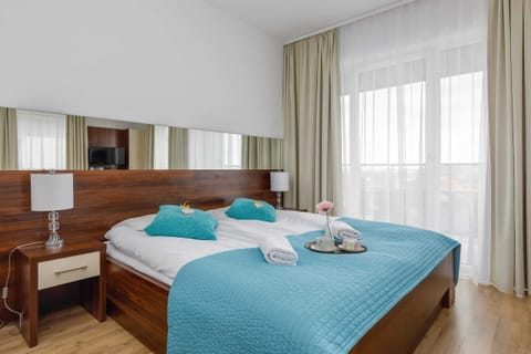 A cozy bedroom featuring a double bed with blue bedding, bedside tables, and large windows with white curtains.