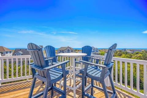 This elevated rooftop deck offers sweeping views of the ocean and surrounding coastal community, featuring a cozy seating setup with Adirondack-style chairs positioned around a small table.