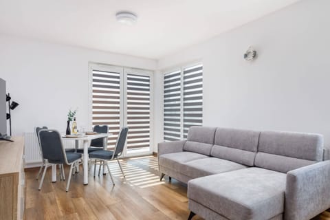  A cozy living room with a comfortable gray sofa, a TV, and a dining area. The space is bright with natural light coming from large windows.