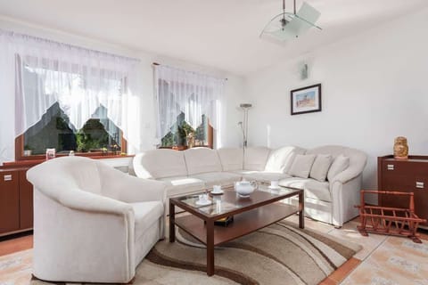 A cozy living room with a white sofa set, armchairs, and a coffee table on a patterned rug near large windows.