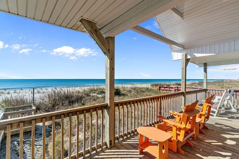 Covered oceanfront deck with bright Adirondack chairs, wood railing, and panoramic views over dunes to the Atlantic Ocean, perfect for relaxing in this Oak Island, NC beachfront home.