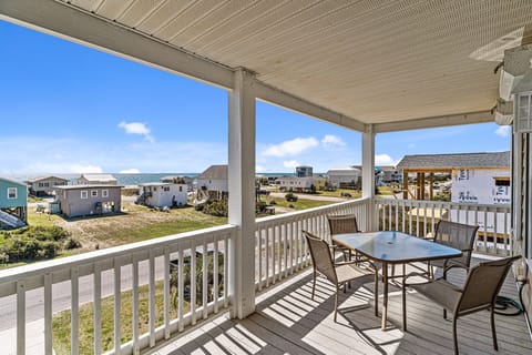 Covered deck with outdoor dining table, coastal views, white railings, and partial ocean views overlooking beach homes. Relax outdoors and enjoy the sea breeze at this Oak Island, NC beach retreat.