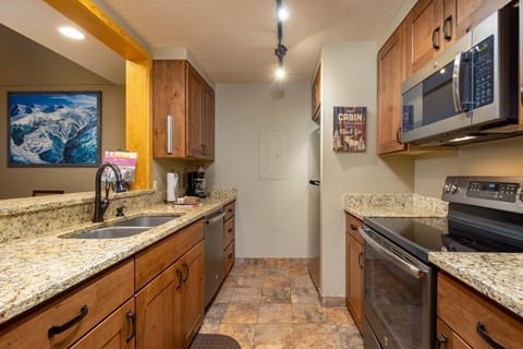A compact kitchen with wooden cabinets, granite countertops, stainless steel appliances, and tile flooring. A framed mountain scenery picture hangs on the left wall, and a cabin sign on the right wall.