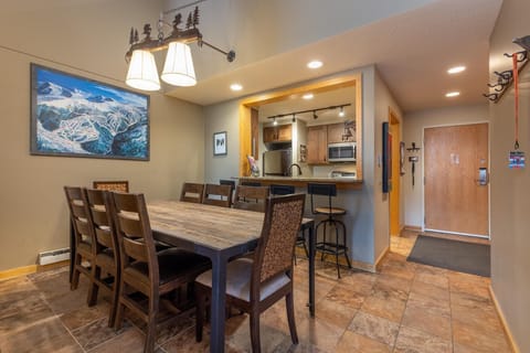 A dining room with a wooden table and six chairs is adjacent to a kitchen with an open counter, two bar stools, and modern appliances. A painting and pendant lights decorate the space.