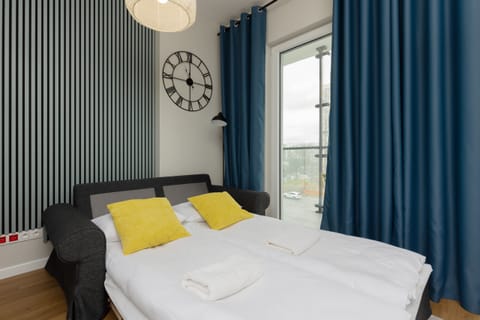 A cozy studio featuring a double bed with white and yellow bedding, framed by large windows with blue curtains for natural lighting.