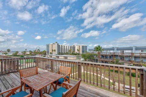 Wooden deck with turquoise-cushioned chairs overlooking coastal development and palm trees under bright blue sky.