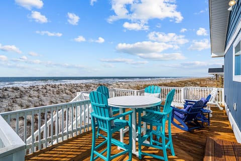 Spacious oceanfront deck in Oak Island featuring turquoise Adirondack chairs, round outdoor dining table, white railings, and panoramic views of sandy dunes and rolling Atlantic waves just steps away.