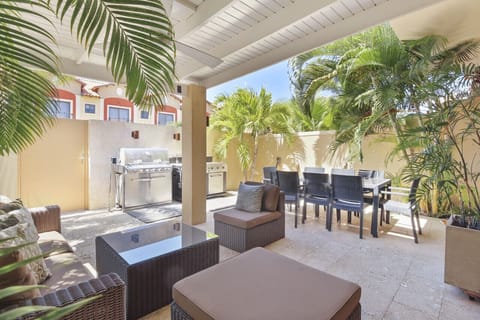 Tropical outdoor patio featuring covered wicker seating, a ceiling fan, outdoor dining setup, stainless steel BBQ grill, and private palm garden.