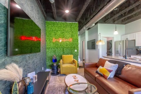 Stylish open living space with a green moss wall, neon signs, leather sofa, and yellow accent chairs flowing into the kitchen
