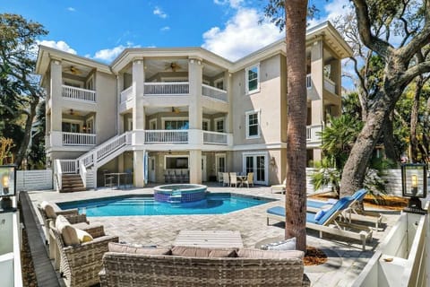 A stunning three-story vacation rental featuring a pristine pool, hot tub, and multiple covered balconies surrounded by mature palm trees.