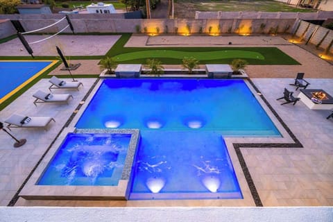 Aerial view of the property's luxurious pool area at dusk, featuring illuminated blue waters, spa section, putting green, and modern outdoor entertaining spaces surrounded by contemporary concrete walls with ambient lighting.