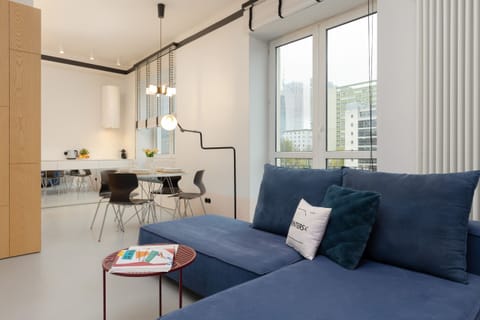 Spacious living room with a modern blue sofa, minimalist decor, and large windows letting in natural light.
