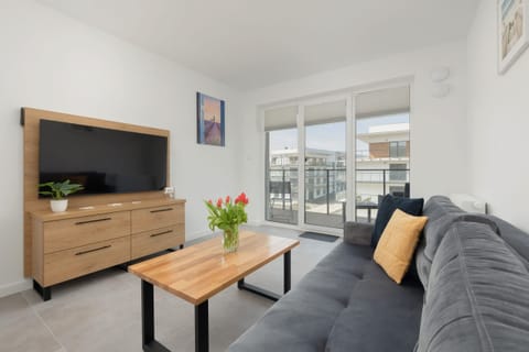 A modern living room with a cozy gray sofa, a wooden coffee table, and a TV stand, bathed in natural light from large windows.

