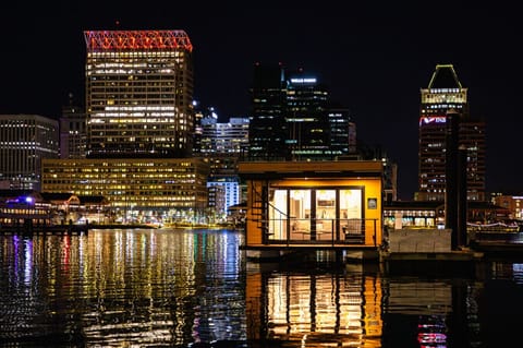 Bay Escape glowing on the Inner Harbor at dusk, framed by the Baltimore skyline with the Royal Sonesta, Wells Fargo, and PNB towers reflecting across the water.