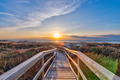 Wooden boardwalk leads through coastal dunes to the beach, with a stunning sunset over the ocean creating a peaceful pathway to the shore.