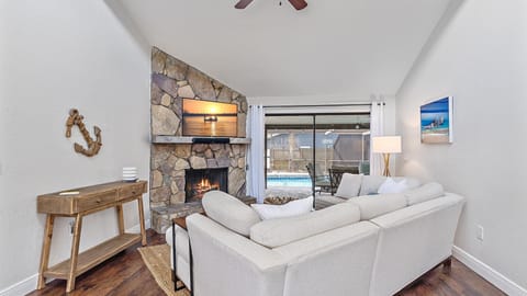 Living room with a stone fireplace, TV mounted above, white sectional sofa, wooden console table, wall decorations, and large sliding glass doors opening to a view of a swimming pool outside.