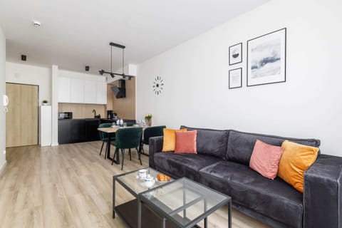 A bright living room with a black sofa, decorated with vibrant cushions, a coffee table, and a large window.