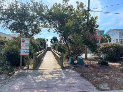 Wooden boardwalk leads through a tree-lined residential area to beach access, with coastal homes visible along the pathway.