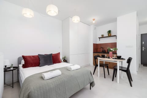 Bright studio apartment with a double bed, red accent pillows, a dining table, and modern lighting.
