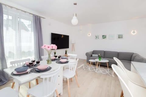 A cozy dining area with a round table set with pink flowers and black plates, placed in a bright, airy room. A television and sofa can be seen in the background.