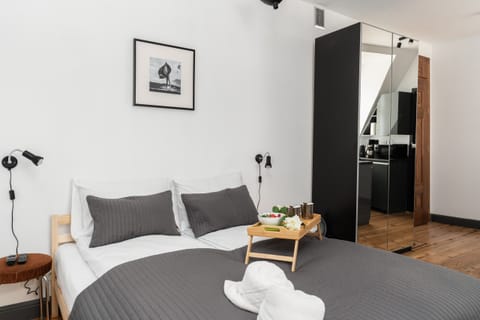 A cozy bedroom featuring a double bed with grey bedding, neatly folded towels, and a bedside table with a decorative lamp.
