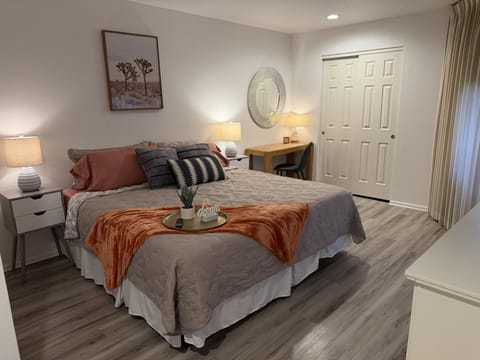 Queen bed with layered pillows and rust throw, flanked by white nightstands and lamps on vinyl plank flooring.