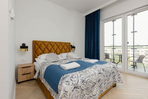 A cozy bedroom featuring a double bed with blue bedding, a mustard-colored headboard, and natural light from large windows.

