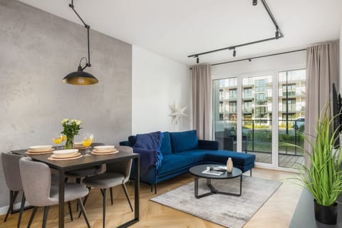 A modern, open-plan living room with a blue sofa, a dining table with chairs, and a large window letting in natural light.