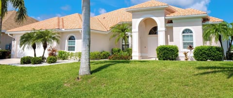 Green lawn and palm trees frame the villa’s soft beige stucco exterior and terracotta tile roof under a clear blue sky.