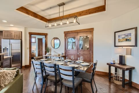 Modern dining area with a wooden table set for eight, metal chairs, and a contemporary chandelier. Adjacent kitchen visible in the background, wooden doors, wall art, and a side table with a lamp.