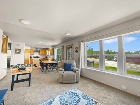 A living room with beige carpet, a recliner, foosball table, wooden dining table, and large windows showing an outdoor scene. The background includes a kitchen with wooden cabinets and stainless-steel appliances.